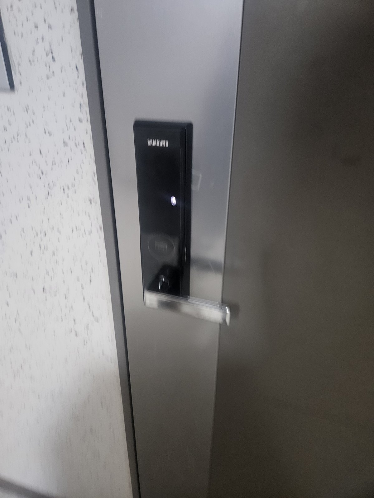 만안구 안양1동 출장 — 은색 문에 달린 검고 길쭉한 레버형 도어락, 가운데 작은 흰 불빛과 흔들려 번진 은색 손잡이